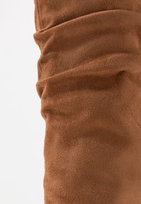 Tejido de ante marrón con textura suave y costuras visibles, mostrando pliegues y arrugas sobre un fondo blanco liso.