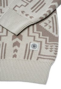 Beige gestrickter Pullover mit hellbraunen geometrischen Mustern und einem kleinen weißen Etikett mit der Aufschrift „EHRLICHKEIT REGIEREN“ nahe dem gerippten Saum.