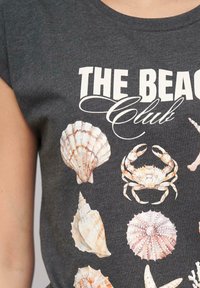 T-paita tummanharmaana, jossa on "THE BEACH CLUB" -grafiikka, jossa on erilaisia ystävällisiä simpukoita ja rapu vaaleissa väreissä. Lyhyt hiha.