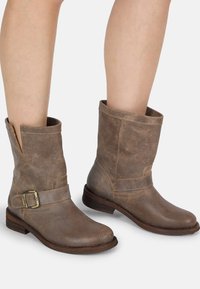 Botas de cuero marrón hasta el tobillo con un acabado texturizado, puntera circular y una correa ancha con una hebilla de metal. Diseño ligeramente caído.