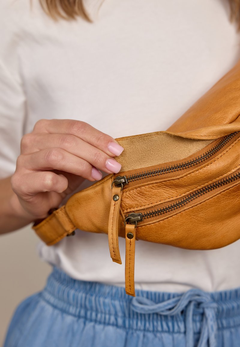 Hand with pink nails opening tan leather waist bag with two zippered compartments, worn over white shirt and blue pants.