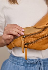 Ranka su rožiniais nagais atidaro šviesiai rudos odos juosmens maišą su dviem užtrauktukais, dėvima ant baltos marškinės ir mėlynų kelnių.