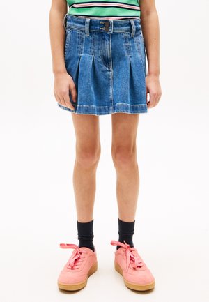 Dziecko ubrane w niebieską plisowaną spódnicę dżinsową, czarne skarpetki i różowe trampki stoi na białym tle.