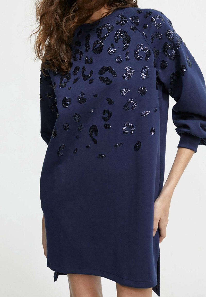 Femme portant une robe bleu marine à manches longues avec un imprimé léopard en sequins noirs, les mains détendues le long du corps, sur un fond clair uni.