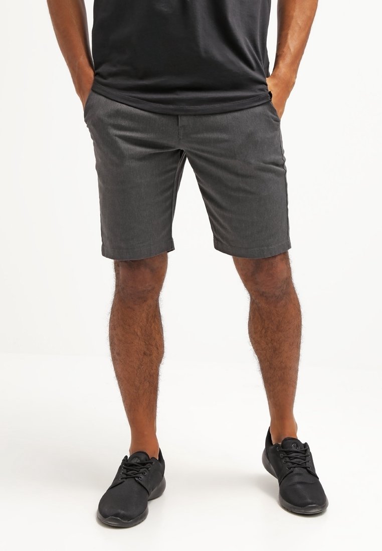 Shorts gris en tissu léger, avec un devant plat et deux poches latérales, associés à des chaussures de sport noires.