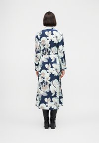 Γυναίκα με σκούρα καρέ μαλλιά που φορά φόρεμα μακρύ με λουλούδια σε μπλε ναυτικό, μπεζ και μέντα, με ζώνη στη μέση, συνδυασμένο με μαύρα ψηλοτάκουνά μποτάκια, όρθια με την πλάτη στραμμένη.