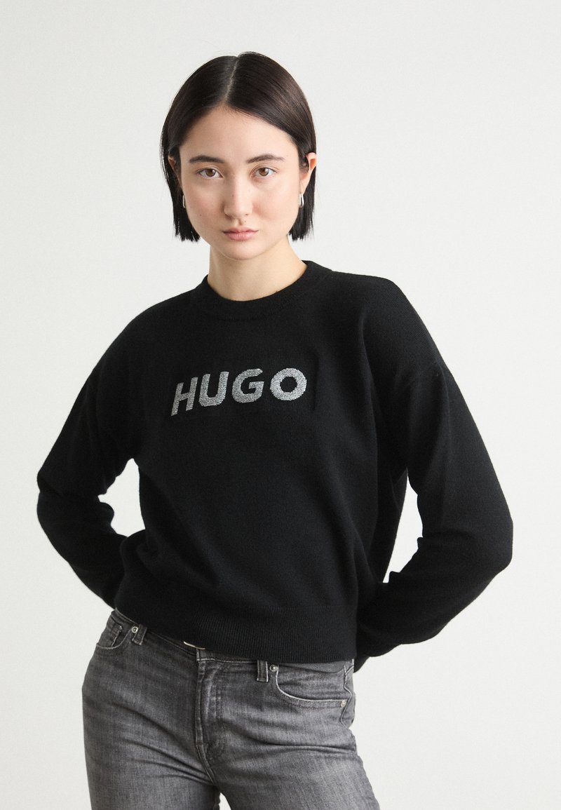 Zwarte gebreide trui met een ronde hals en het woord "HUGO" in metallic zilveren letters over de borst. Draagt het met grijze spijkerbroek.