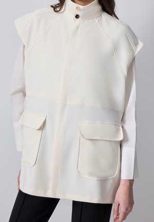 Femme portant un gilet blanc sans manches à col montant avec deux grandes poches avant, sur une chemise à manches longues et un pantalon noir.