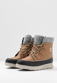 Ein Paar braune und schwarze, isolierte Winterstiefel mit grauem Fleece-Futter und beigen Sohlen auf einer weißen Oberfläche und im Hintergrund.