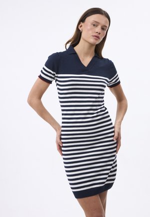 Kobieta w granatowo-białej sukience z krótkimi rękawami, poziome paski, z kołnierzykiem, stojąca z rękami na biodrach na białym tle.