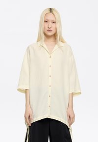 Camicia corta gialla pallido con maniche corte e bottone frontale, caratterizzata da un colletto e dettagli a bordo arricciato con lacci regolabili sui lati.