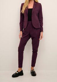 Paarse blazer en bijpassende broek gemaakt van gladde stof. De blazer heeft een enkele knoop en revers. Zwarte schoenen met een metalen accent.