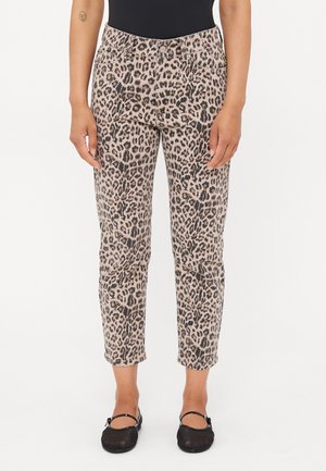 Personne portant un pantalon beige et noir imprimé léopard, associé à des ballerines noires, debout avec les bras détendus le long du corps sur un fond uni.