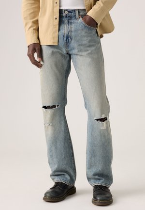 Personne portant un jean déchiré bleu clair, des chaussures noires, une chemise beige boutonnée et un sous-vêtement blanc, debout avec une main dans la poche.