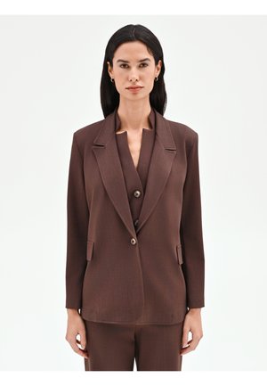 Donna con capelli neri lisci indossa un blazer marrone sopra un gilet abbottonato e pantaloni abbinati, in piedi contro uno sfondo bianco semplice.