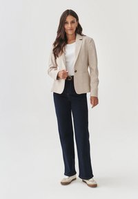 Beige saka na míru s klopovými límci, zapínání na knoflíky, v kombinaci s tmavě modrými džíny rovného střihu a bílými teniskami s hnědými detaily.