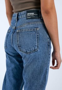 Jeans in denim blu a vita alta con lavaggio sbiadito, cuciture visibili e due tasche posteriori. Presenta un'etichetta nera in vita.