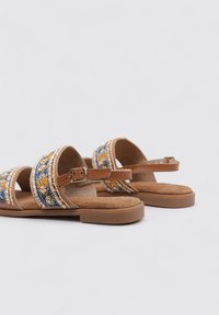 Sandalias marrones con tres correas bordadas que presentan patrones en azul, amarillo y blanco. Cierre de hebilla y suela plana y texturizada.