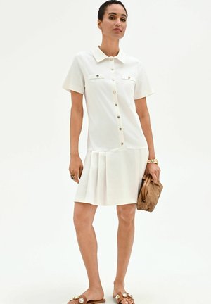 Femme portant une robe blanche à manches courtes boutonnée avec des plis, tenant un sac à main tissé marron et portant des sandales plates ornées de pierres décoratives.