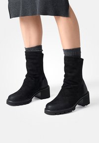 Bottes chevilles en suede noir avec des semelles en caoutchouc texturé, des talons carrés et un design décontracté. Bordées d'un détail en chaussette tricotée grise.