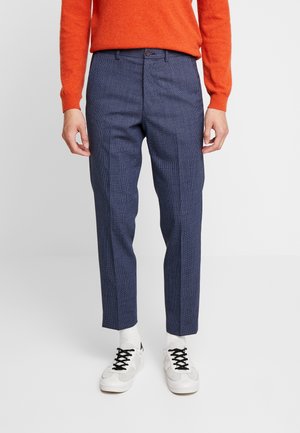 Mand iført teksturerede blå skræddersyede bukser, hvide sokker, hvide sneakers med sorte snørebånd og en orange langærmet sweater, stående på en ensfarvet baggrund.