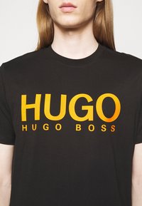 Camiseta de algodón negra con texto en degradado naranja audaz, "HUGO HUGO BOSS," con mangas cortas y escote redondo.
