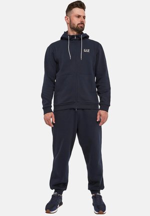 Navy zip-up hoodie with drawstrings, front pockets, and logo, paired with matching joggers and dark athletic shoes. Casual athletic wear.