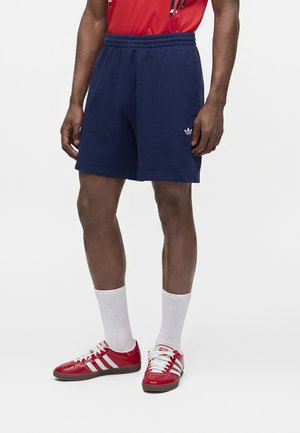 Muž nosiaci námornícke modré kraťasy s logom Adidas, biele ponožky, červené tenisky Adidas a červené tričko, stojaci na jednofarebnom pozadí.