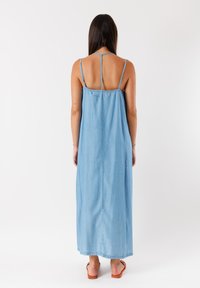 Abito lungo in denim blu con spalline sottili, vestibilità comoda, lunghezza a metà polpaccio e una texture liscia. Nessun motivo o decorazione visibile.