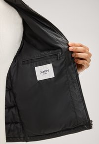Schwarze Jackeninnenseite mit glattem Futter, ausgestattet mit einer Reißverschlusstasche und einem Etikett, auf dem "JOOP! JEANS" steht. Die Textur wirkt weich und elegant.