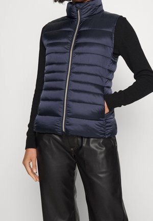 Femme portant une veste matelassée sans manches bleu marine avec fermeture éclair devant, un chemisier noir à manches longues et un pantalon en cuir noir sur fond blanc.
