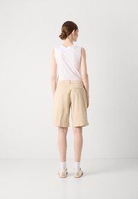 Bermudas en coton beige avec une coupe décontractée, possédant des ourlets retroussés et une poche au dos, accompagnées d'un haut sans manches blanc et de chaussures beiges.