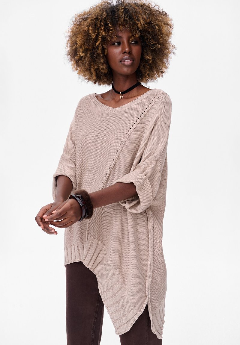 Beige oversized gebreid sweater met een ronde halslijn, zijsplitten en een geribbelde zoom. Draag over bruine broek. Zachte textuur.