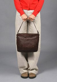 Bolso de cuero tejido marrón con cierre de cremallera y una sola correa, sostenido por una persona con un cárdigan rojo y pantalones de color claro.