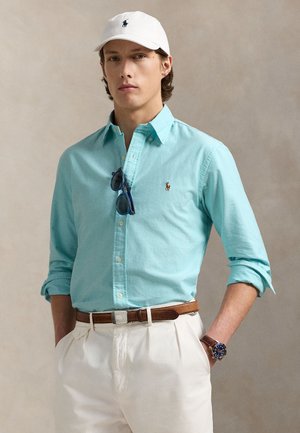 Jeune homme portant une casquette blanche, une chemise bleu clair boutonnée avec des lunettes de soleil accrochées au col, un pantalon blanc, une ceinture marron et une montre-bracelet.