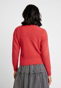 Pull rouge en maille avec une texture côtelée et un empiècement dos arrondi, assorti à une jupe à carreaux noir et blanc.