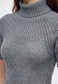 Pull gris côtelé à col roulé avec manches courtes porté par une personne montrant le cou et le haut du torse.