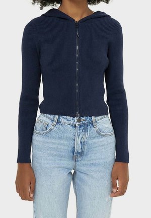 Persoon draagt een marineblauwe geribbelde capuchontrui met rits en lichtblauwe high-waisted jeans, staand met de armen ontspannen langs het lichaam.
