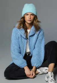 Cardigan bleu tricoté avec des manches surdimensionnées et une capuche, associé à un bonnet côtelé bleu clair. Le mannequin porte un haut blanc et un pantalon foncé.