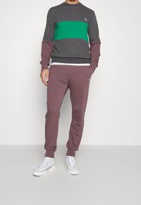 Sudadera gris con una raya verde, combinada con pantalones de chándal color burdeos. Ambas prendas están confeccionadas con una tela suave. Unas zapatillas blancas completan el conjunto.