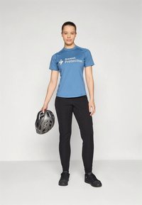 Camisola azul de manga curta com o logotipo "Sweet Protection", calças pretas de corte justo e um capacete cinzento segurado numa mão.