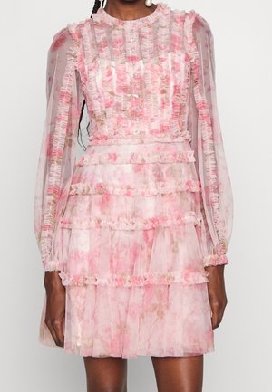 Kvinde iført en gennemsigtig lyserød blomsterkjole med flæsekantede lodrette striber, lange pufærmer, knappet front og lagdelt skørt.