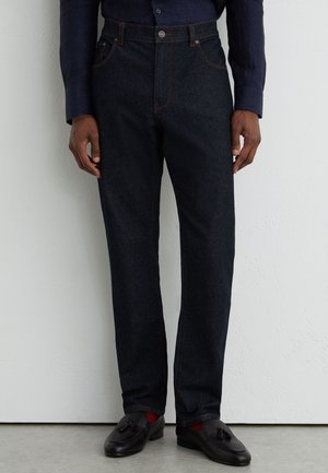 Mand iført mørkeblå jeans, sorte frynsemokkasiner og en mørk langærmet skjorte, der står op ad en hvid væg.