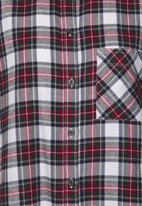 Camisa de franela a cuadros en rojo, negro y blanco con botón en la parte delantera, un bolsillo en el pecho y una textura que muestra varios patrones de cuadros.