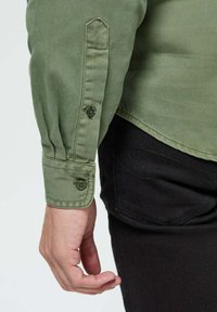 Chemise olive à col boutonné avec un poignet ajusté, dotée de deux boutons et d'un ourlet droit, assortie à un pantalon noir.