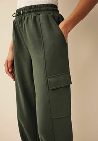 Ciemnozielone spodnie dresowe wykonane z miękkiego materiału. Posiadają elastyczną talię, boczne kieszenie oraz kieszeń cargo na udzie. Gładka faktura.