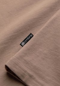 Ruskea puuvillapaita, jossa on taitettu hiha ja musta etiketti, jossa on "ombre"-logo. Su smooth tekstuuri ja hienovarainen ompeludetail.