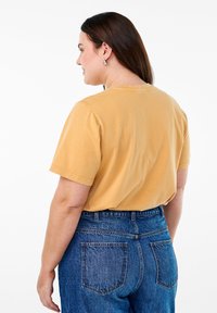 Camiseta de algodón color mostaza con mangas cortas, cuello redondo y un corte relajado, combinada con unos vaqueros de talle alto en denim azul.