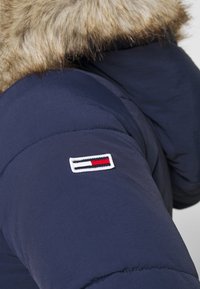 Mörkblå jacka med en plysch, ljusbrun pälskapuschong och en liten broderad logotyp med röda, vita och svarta ränder på ärmen.