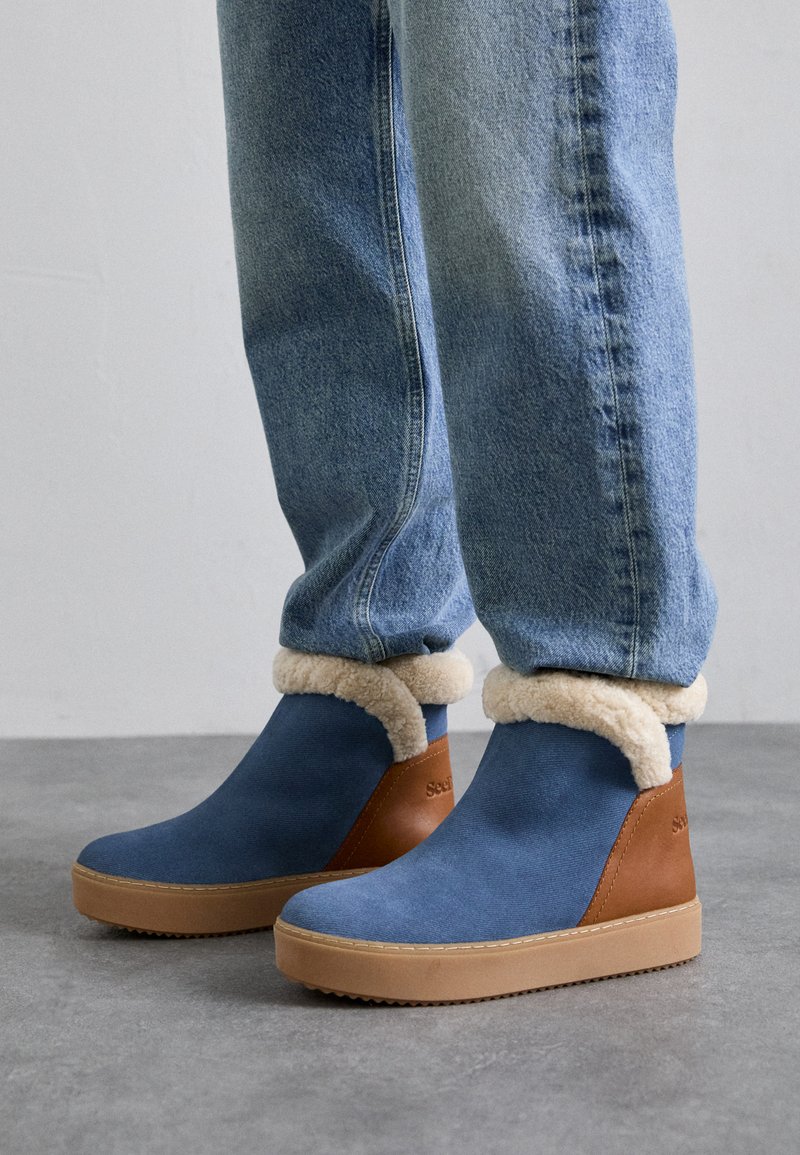 Bottines bleu et marron avec doublure en fourrure beige portées avec un jean bleu clair sur un sol gris.